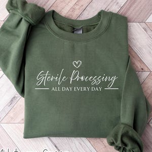 May include: A military green crewneck sweatshirt featuring white screen-printed text that reads Sterile Processing ALL DAY EVERY DAY with a small heart icon above the text. The garment has long sleeves with ribbed cuffs and a ribbed neckline.