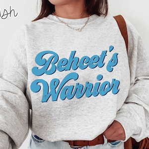 May include: A light gray sweatshirt with the text "Beheet's Warrior" in a blue, retro font. The sweatshirt has long sleeves and a crew neck. The word "Ash" is written in the upper left corner.