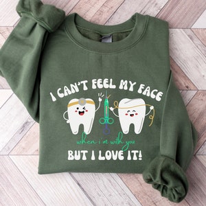 May include: Green sweatshirt with the text "I CAN'T FEEL MY FACE when I'm with you BUT I LOVE IT!". The design features two cartoon teeth, a syringe, and dental floss. The sweatshirt is folded on a wooden surface.
