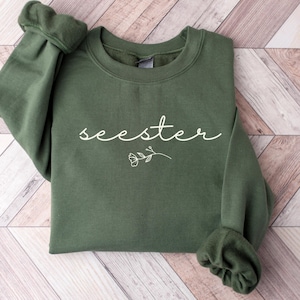 May include: A dark green sweatshirt with the word "seester" written in white script with a small flower graphic.