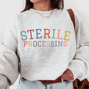 May include: A light gray heathered crewneck sweatshirt featuring the text STERILE PROCESSING printed in a multi-colored collegiate font across the chest. The letters alternate between blue, red, orange, green, pink, and yellow hues.