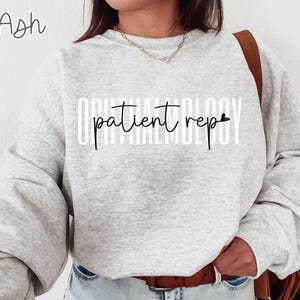 May include: Light gray sweatshirt with the words "OPHTHALMOLOGY patient rep" printed in white and black. The word "Ash" is written in the upper left corner. The sweatshirt has long sleeves and a crew neck.