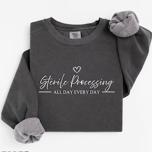 May include: A charcoal gray crewneck sweatshirt featuring the white text Sterile Processing ALL DAY EVERY DAY with a small heart icon above the text. The garment has long sleeves with ribbed cuffs and a Comfort Colors brand label at the neckline.