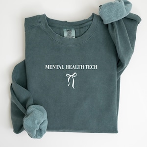 May include: A teal-colored sweatshirt with the text "MENTAL HEALTH TECH" in white, along with a white bow graphic. The sleeves are folded, and the garment appears to be made of a soft material.