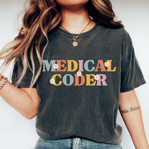 May include: A charcoal gray short-sleeve t-shirt featuring the text MEDICAL CODER in a multi-colored retro font. The letters are accented with small white daisy graphics. The shirt has a crew neckline and a relaxed fit, shown in the color pepper.