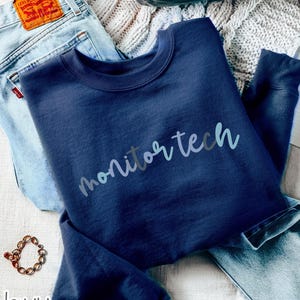 May include: A navy blue long-sleeve sweatshirt featuring the text "monitor tech" in a light blue script font. The garment is positioned alongside light blue denim jeans and a gold-toned chain bracelet on a textured white surface.
