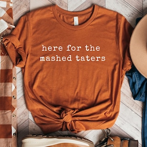 May include: A rust-colored t-shirt with the text "here for the mashed taters" in white lettering.