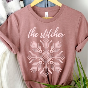May include: A mauve t-shirt with the text "the stitcher" in pink lettering and a pink cross-stitch design.