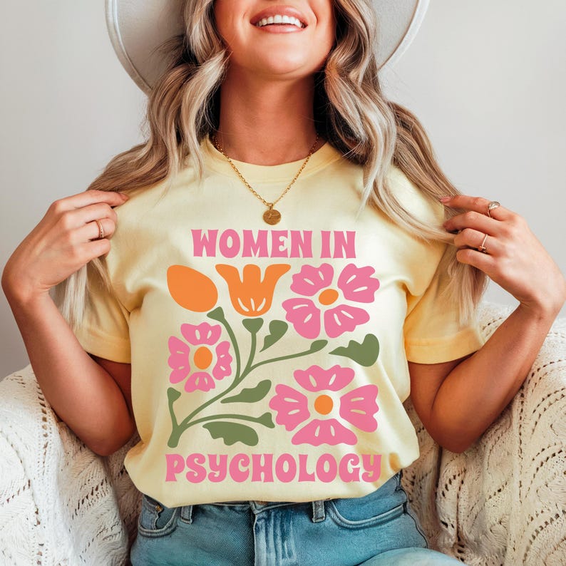 K&ouml;nnte beinhalten: Ein gelbes T-Shirt mit einem Blumendesign und dem Text "Women in Psychology" in rosa Buchstaben.
