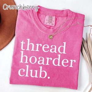 May include: A bright pink t-shirt with the words "thread hoarder club." printed in white. The shirt has a crew neck and short sleeves. A gold necklace with a small pendant is draped across the shirt.