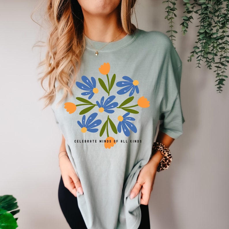 May include: A light blue t-shirt featuring a floral design in shades of blue, green, and orange. The text "Celebrate Minds of All Kinds" is printed below the design.