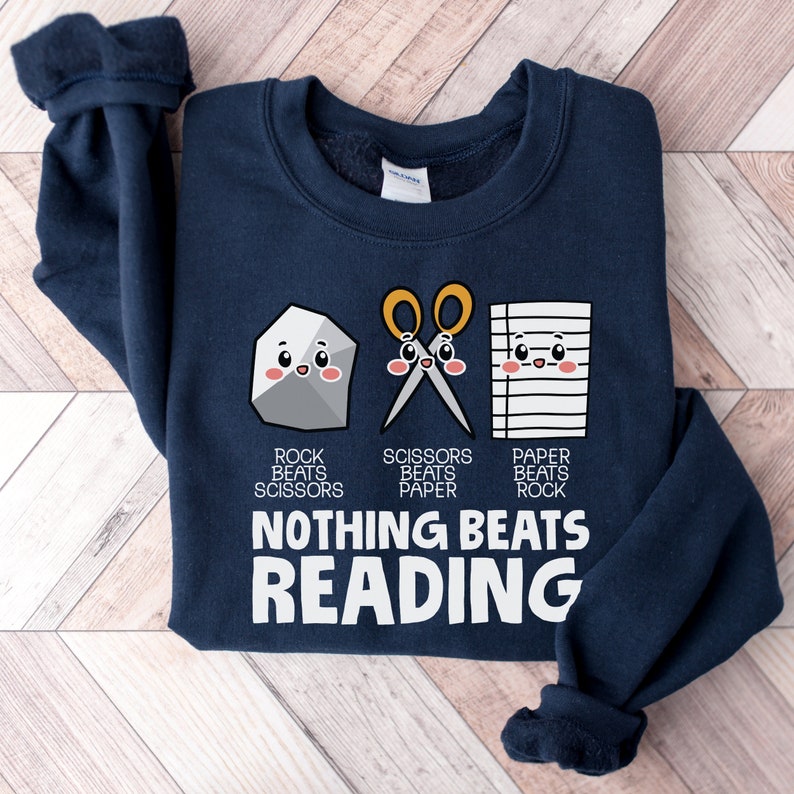 Puede incluir: Una sudadera azul marino con un gr&aacute;fico blanco de una roca, tijeras y papel con caras. El texto dice "Rock beats Scissors, Scissors beats Paper, Paper beats Rock, Nothing beats Reading."