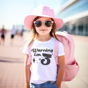 May include: A young girl wearing a white t-shirt with the text "Warning I'm 3" in black, a pink hat, and pink sunglasses. She is carrying a pink backpack.