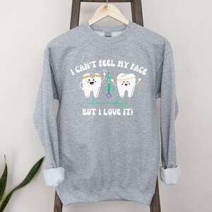 May include: A grey sweatshirt featuring the text "I CAN'T FEEL MY FACE" above two cartoon teeth characters. One tooth wears a headband and holds a syringe, the other holds floss. The text "who cares as long as it's with you BUT I LOVE IT!" is below.