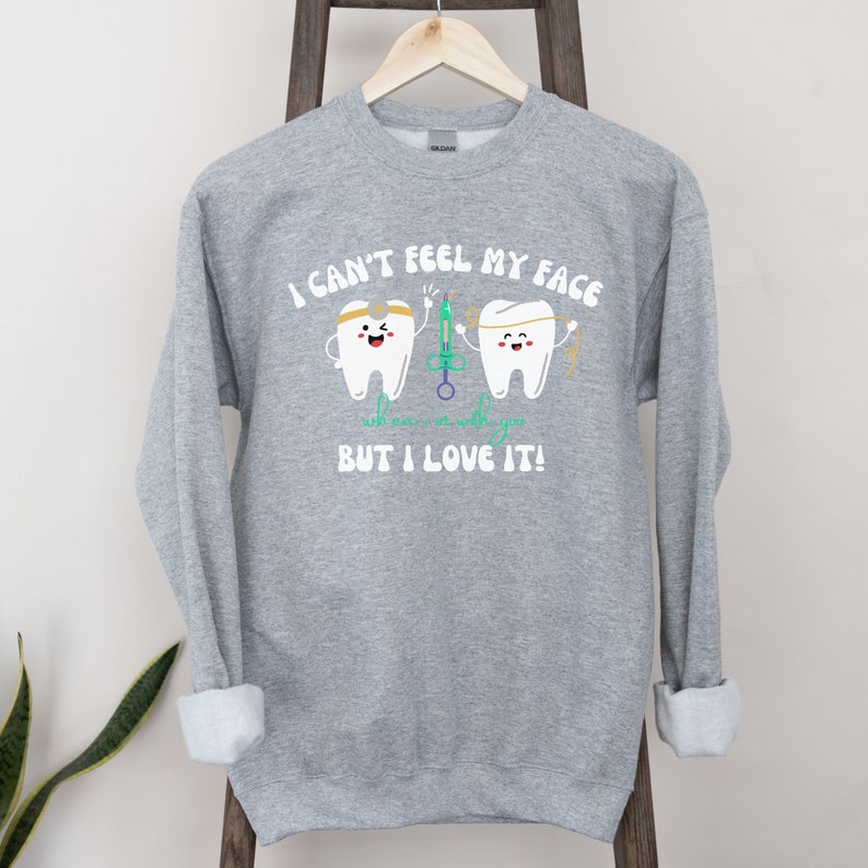 May include: A grey sweatshirt featuring the text "I CAN'T FEEL MY FACE" above two cartoon teeth characters. One tooth wears a headband and holds a syringe, the other holds floss. The text "who cares as long as it's with you BUT I LOVE IT!" is below.