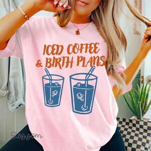 Puede incluir: Camiseta rosa claro con el texto "ICED COFFEE & BIRTH PLANS" en letras marrones. Debajo, dos vasos azules con contorno de caf&eacute; helado con pajitas. La camiseta tiene mangas cortas y un corte holgado.