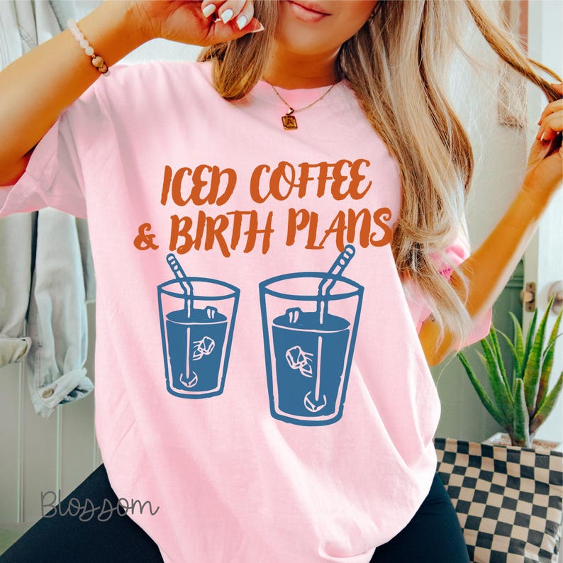 Puede incluir: Camiseta rosa claro con el texto "ICED COFFEE & BIRTH PLANS" en letras marrones. Debajo, dos vasos azules con contorno de caf&eacute; helado con pajitas. La camiseta tiene mangas cortas y un corte holgado.