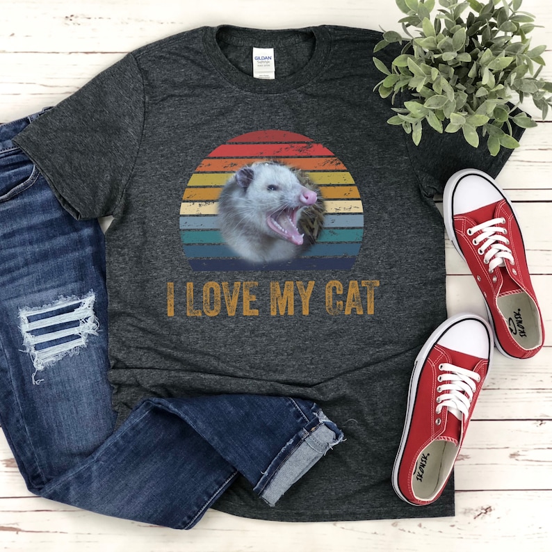 Puede incluir: Una camiseta gris oscuro con un gr&aacute;fico de puesta de sol vintage que presenta un opossum. El texto "I LOVE MY CAT" est&aacute; impreso debajo del gr&aacute;fico.