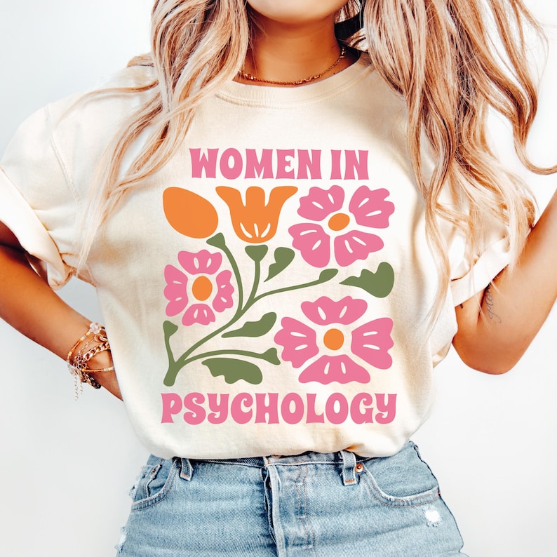 K&ouml;nnte beinhalten: Ein wei&szlig;es T-Shirt mit dem Text "Women in Psychology" in orangefarbenen und rosafarbenen Buchstaben. Der Text ist von einem Blumendesign mit rosa und orangefarbenen Blumen umgeben.