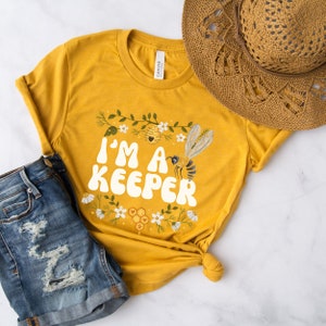 Puede incluir: Una camiseta amarilla con un dise&ntilde;o de abeja y flores. El texto en la camiseta dice "I'm a Keeper".