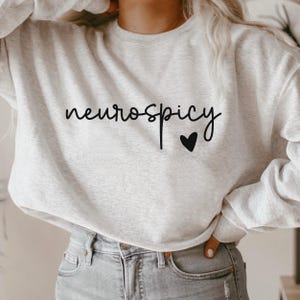Op de afbeelding: Een grijs sweatshirt met de tekst "neurospicy" en een klein zwart hartje geprint op de voorkant.