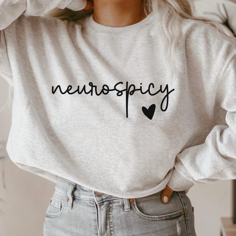 Op de afbeelding: Een grijs sweatshirt met de tekst "neurospicy" en een klein zwart hartje geprint op de voorkant.