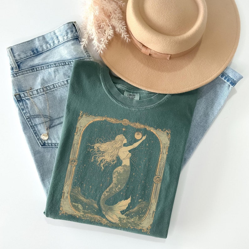 Może przedstawiać: Zielony T-shirt z vintage ilustracją syreny trzymającej perłę. Syrena jest otoczona gwiazdami i falami. Ilustracja jest oprawiona w złotą ramę.