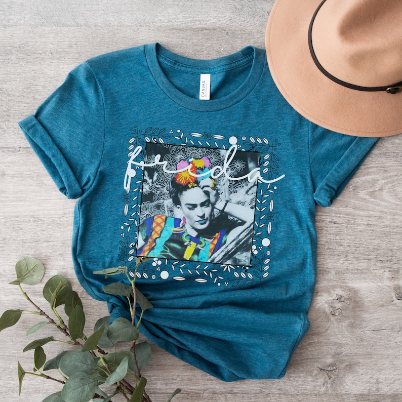 May include: Teal blue t-shirt with a graphic print of Frida Kahlo, a floral border, and the word "frida" written in white cursive.