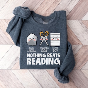 Puede incluir: Una sudadera gris oscura con un gr&aacute;fico de una roca, tijeras y papel con caras. El texto debajo del gr&aacute;fico dice "Rock beats Scissors, Scissors beats Paper, Paper beats Rock, Nothing Beats Reading".