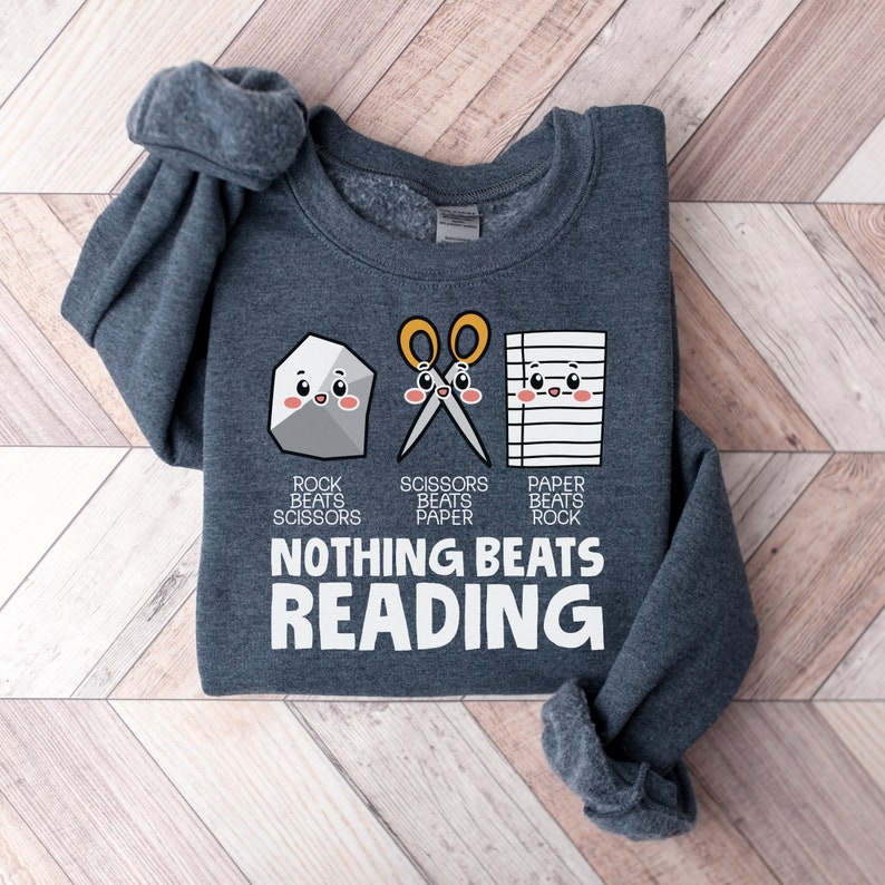 Puede incluir: Una sudadera gris oscura con un gr&aacute;fico de una roca, tijeras y papel con caras. El texto debajo del gr&aacute;fico dice "Rock beats Scissors, Scissors beats Paper, Paper beats Rock, Nothing Beats Reading".
