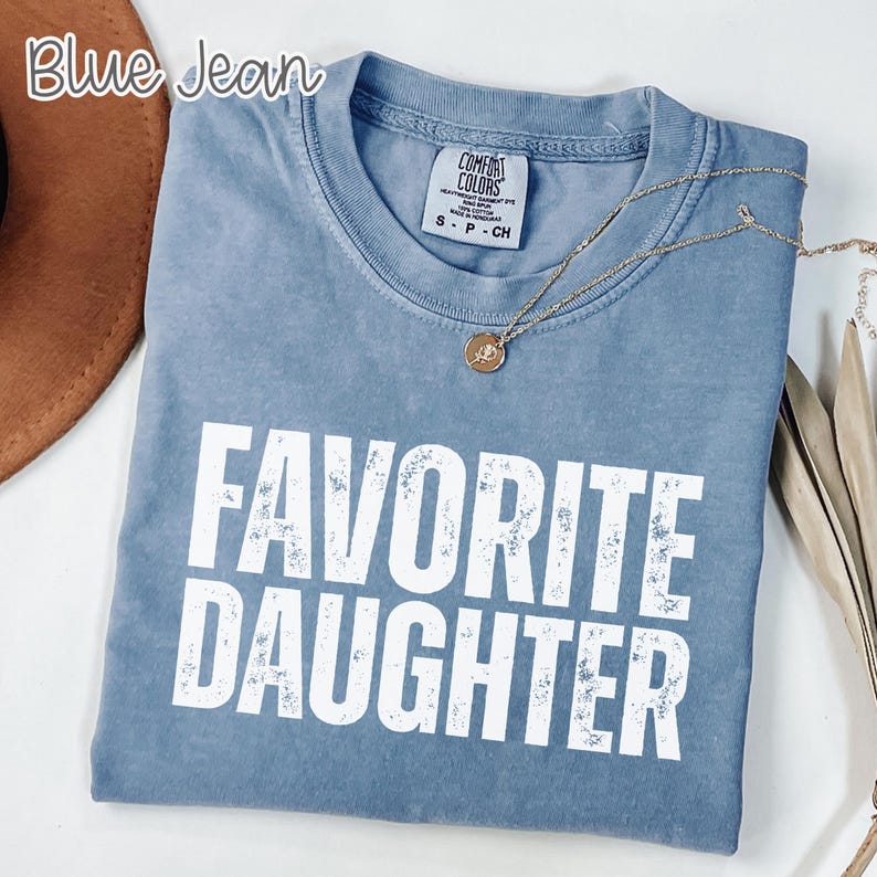 May include: A blue jean coloured short-sleeve t-shirt with a crew neckline and a white graphic print that reads FAVORITE DAUGHTER in a distressed font. The shirt features a Comfort Colors brand label at the inner neck and a gold-toned necklace.