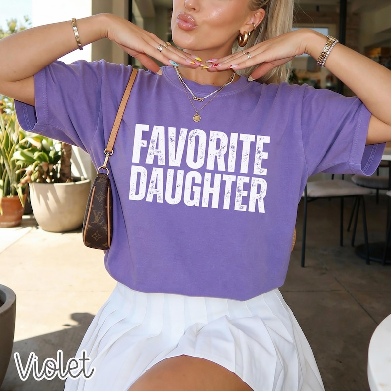 May include: A purple short-sleeved t-shirt featuring the text "FAVORITE DAUGHTER" in white, distressed capital letters across the chest. The shirt has a crew neckline and is paired with a white pleated skirt and a brown patterned shoulder bag.