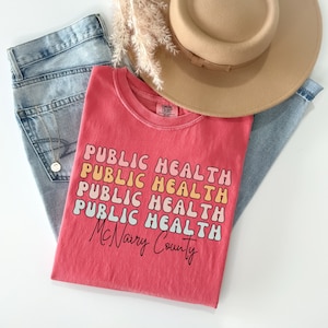 May include: A coral-colored t-shirt with the words "PUBLIC HEALTH" stacked multiple times in a retro font. Below, the text "McNairy County" is written in cursive. The shirt is paired with light-wash jeans, a tan hat, and decorative dried grass.