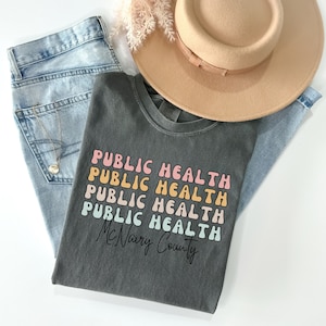 May include: A dark gray t-shirt with the words "PUBLIC HEALTH" stacked in a retro font, with the text "McNairy County" below. The shirt is paired with light blue jeans, a tan hat, and a gold necklace.