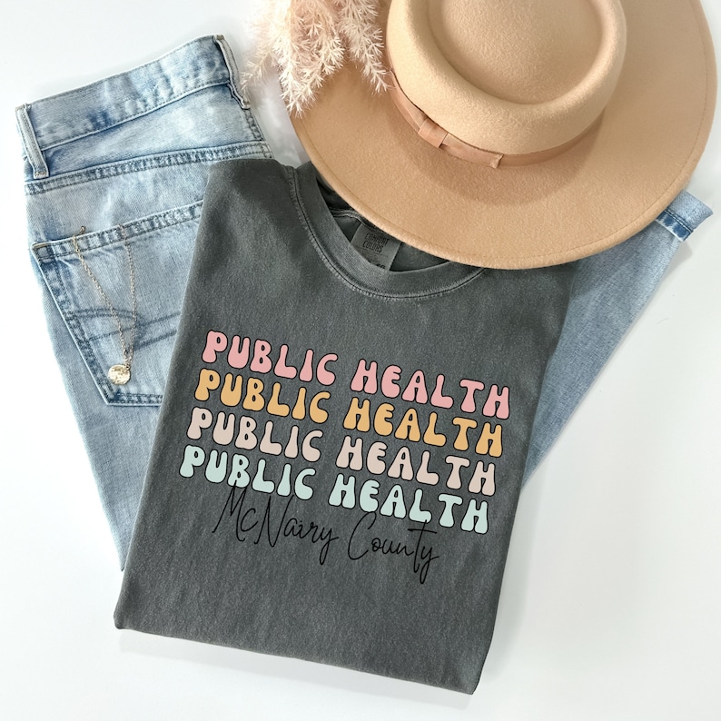 May include: A dark gray t-shirt with the words "PUBLIC HEALTH" stacked in a retro font, with the text "McNairy County" below. The shirt is paired with light blue jeans, a tan hat, and a gold necklace.