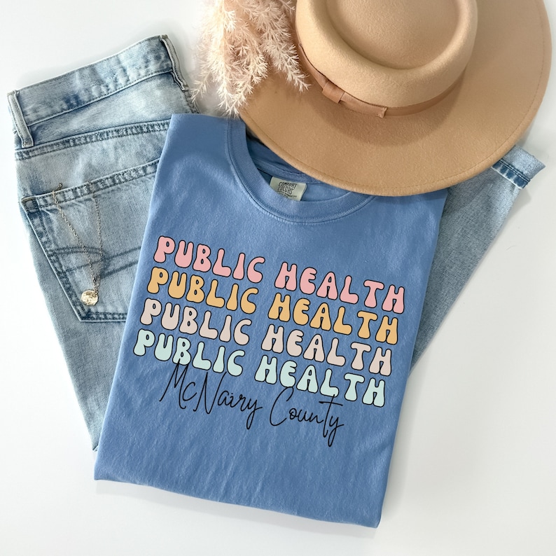 May include: A light blue t-shirt with the words "PUBLIC HEALTH" stacked in a retro font, with "McNairy County" below. The shirt is laid on top of light wash jeans, a tan hat, and a necklace.