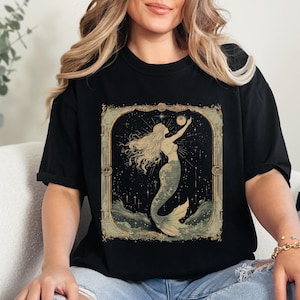 Może przedstawiać: Czarny T-shirt z vintage'ową ilustracją syreny sięgającej księżyca na tle gwieździstego nieba. Ilustracja jest oprawiona w złotą ramkę.