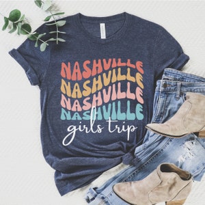 K&ouml;nnte beinhalten: Ein marineblaues T-Shirt mit dem Text "Nashville girls trip" in einem Regenbogen-Farbschema. Der Text ist &uuml;bereinander angeordnet und in einer Retro-Schriftart.
