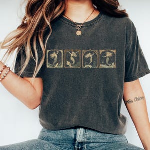 May include: A dark gray t-shirt with a vintage illustration of five mermaids in a row. The mermaids are in different poses, and the illustration is surrounded by a gold frame.