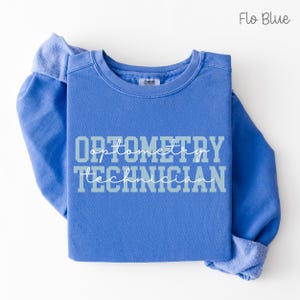 May include: A blue crew neck sweatshirt with the words "OPTOMETRY TECHNICIAN" printed across the front in a two-tone design. The top is a solid blue color, with the text in light blue and white. The sweatshirt is folded.