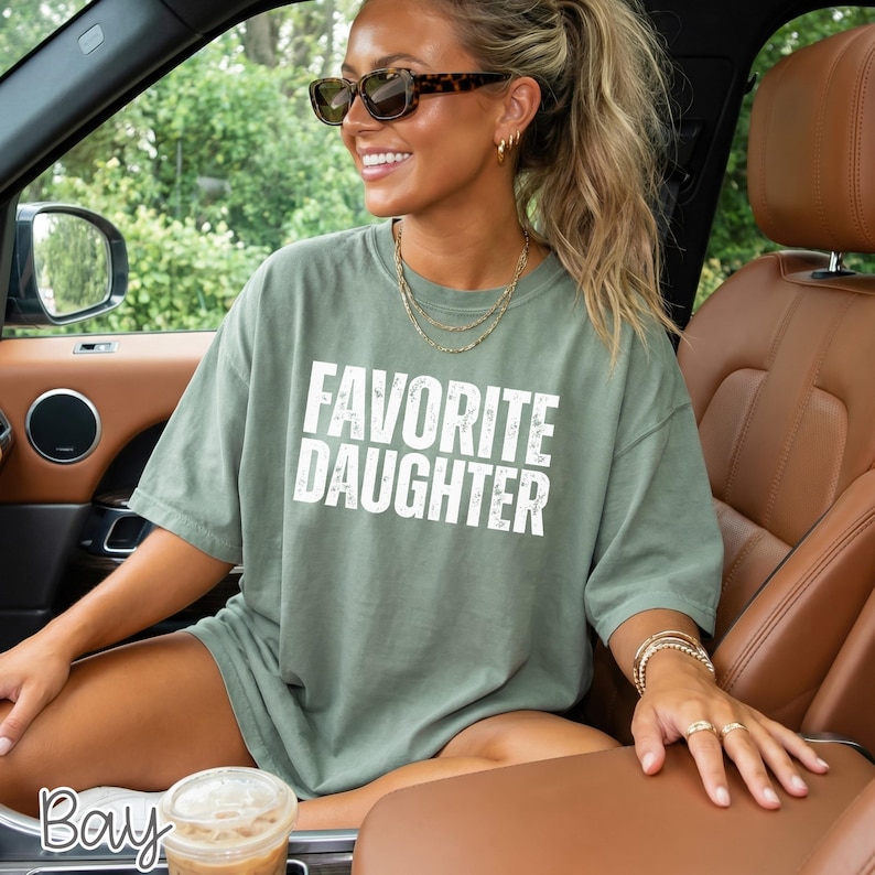 May include: A sage green short-sleeved t-shirt featuring the white distressed text FAVORITE DAUGHTER printed on the chest. The shirt has a crew neckline and a relaxed fit. The wearer is also sporting dark tortoise shell sunglasses.