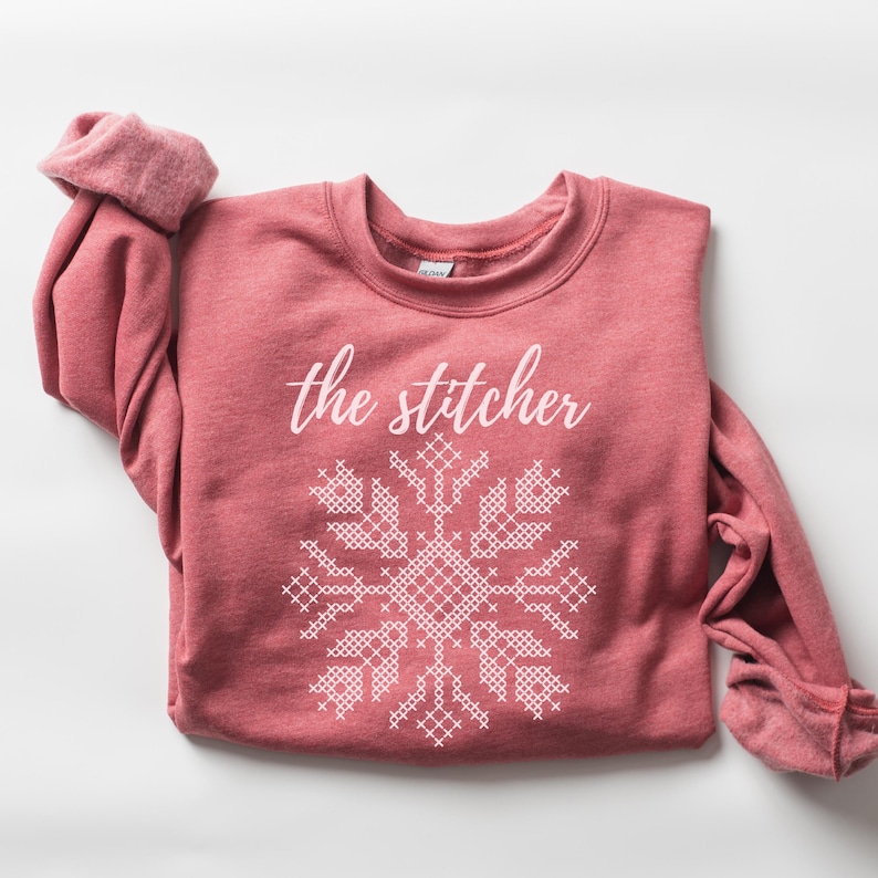 May include: A red-pink sweatshirt with the text "the stitcher" above a white cross-stitch snowflake design. The sleeves are cuffed, and the crew neck is visible. The sweatshirt is made of a soft material.