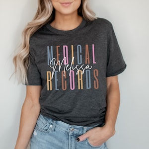 May include: A dark gray t-shirt with the text "MEDICAL Melissa RECORDS" printed in colorful vertical stripes. The text is in a variety of colors, including pink, orange, blue, and brown.