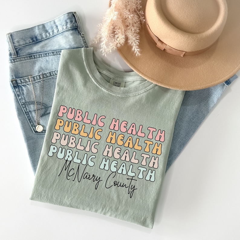 May include: A sage green t-shirt with the words "PUBLIC HEALTH" stacked in a retro font, with "McNairy County" below. The shirt is paired with light wash jeans, a gold necklace, and a tan hat.