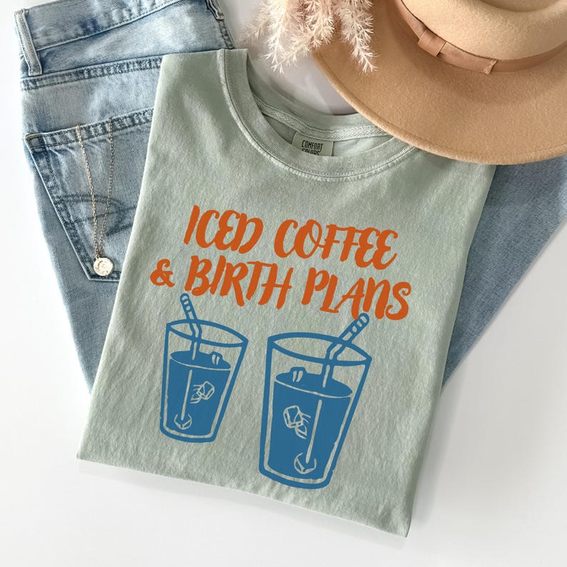 Puede incluir: Una camiseta verde claro con el texto naranja "ICED COFFEE & BIRTH PLANS". Debajo, dos dibujos azules de caf&eacute; helado. La camiseta est&aacute; sobre unos vaqueros azules, un collar y un sombrero beige.