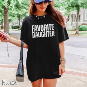 May include: A black short-sleeved t-shirt featuring the white text FAVORITE DAUGHTER printed in a distressed, bold, sans-serif font across the chest. The garment has a crew neckline and a relaxed, loose-fitting silhouette with a straight hemline.