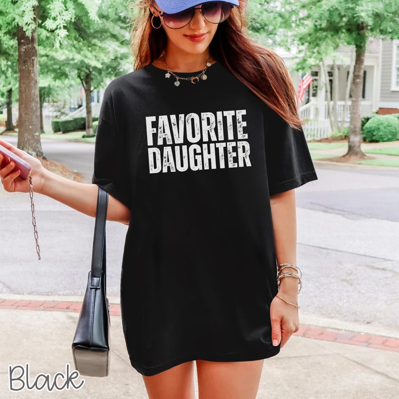 May include: A black short-sleeved t-shirt featuring the white text FAVORITE DAUGHTER printed in a distressed, bold, sans-serif font across the chest. The garment has a crew neckline and a relaxed, loose-fitting silhouette with a straight hemline.
