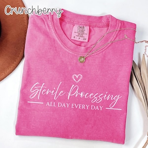 May include: A bright pink short-sleeved t-shirt featuring the white text "Sterile Processing ALL DAY EVERY DAY" with a small heart icon above the text. The shirt has a crew neckline and a Comfort Colors brand label inside the collar area.