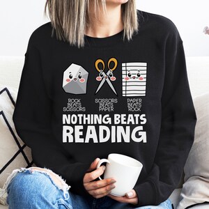 Puede incluir: Sudadera negra con un gr&aacute;fico de piedra, papel, tijeras con el texto "Nada supera la lectura".