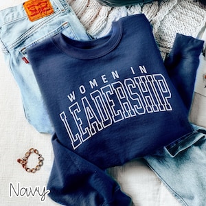 May include: A navy blue crewneck sweatshirt featuring the white text "WOMEN IN LEADERSHIP" in a bold, collegiate-style font. The sweatshirt is positioned alongside light blue denim jeans and a gold-toned chain link bracelet on a white surface.